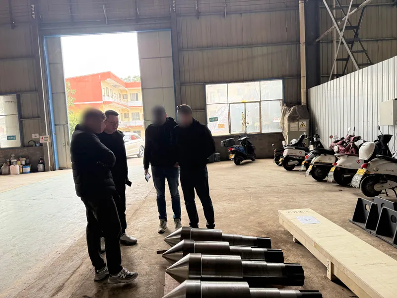 Four men by the shop door beside a row of tapered shafts laid out on the floor.