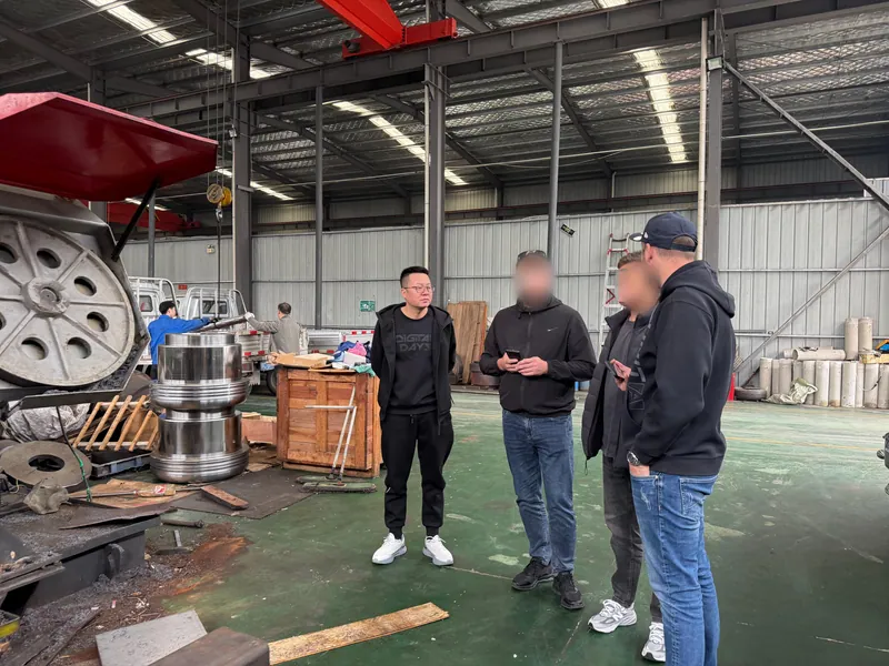A customer beside a finished stepped shaft, talking with our colleague; a vertical lathe chuck and packing case nearby.
